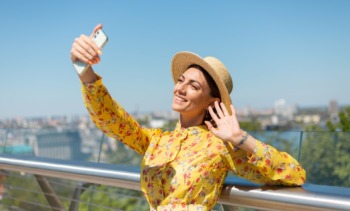 outdoor portrait woman yellow summer dress hat take selfie phone stands bridge with city amazing view min